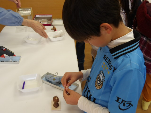 集会室にて、どんぐり工作の先生に木の実を使った蛙のオブジェの作り方を教わりながら工作をする小学生の写真