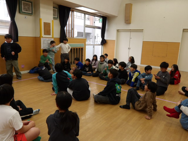 集会室で学年別に並びながら座る小学生と、並びながら座る小学生の前に立ち、ドッジボール大会の進行をする小学生4人の写真