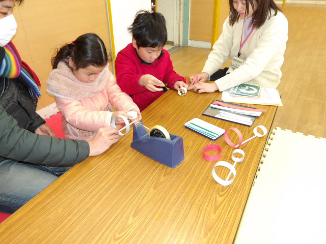 ストローと細く切った画用紙を使い、紙飛行機を作る小学生と幼児親子と職員の写真