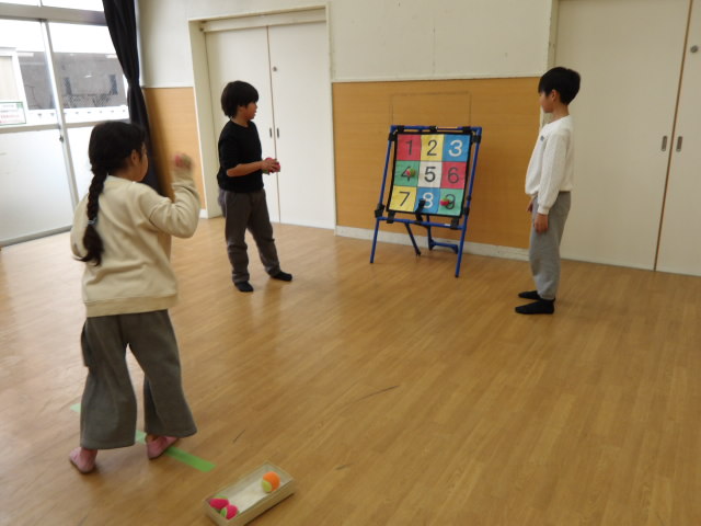 集会室でストラックアウトに挑戦をする女子小学生と、的の両端でボールを回収する男子小学生2人の写真