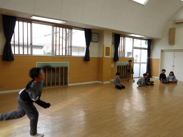 集会室の端からストラックアウトの的に向かって、ボールを投げる小学生とその様子を見ている小学生達の写真