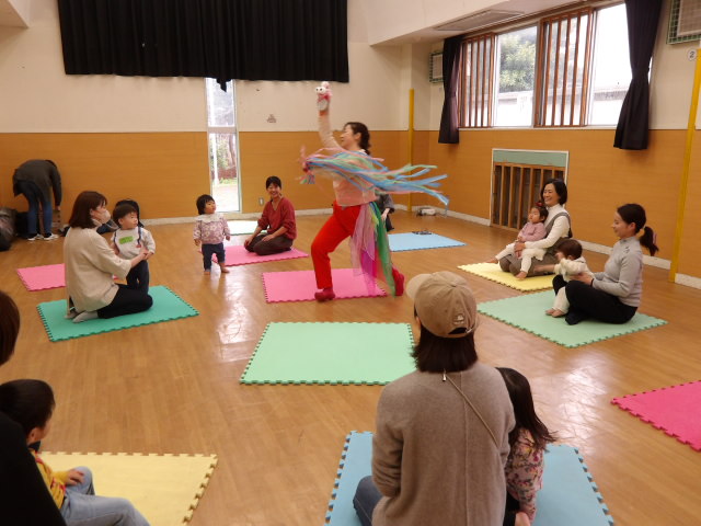 集会室の真ん中でコンテンポラリーダンスを披露しながら自己紹介をする講師と、マットに座りながらパフォーマンスを見る幼児親子達の写真