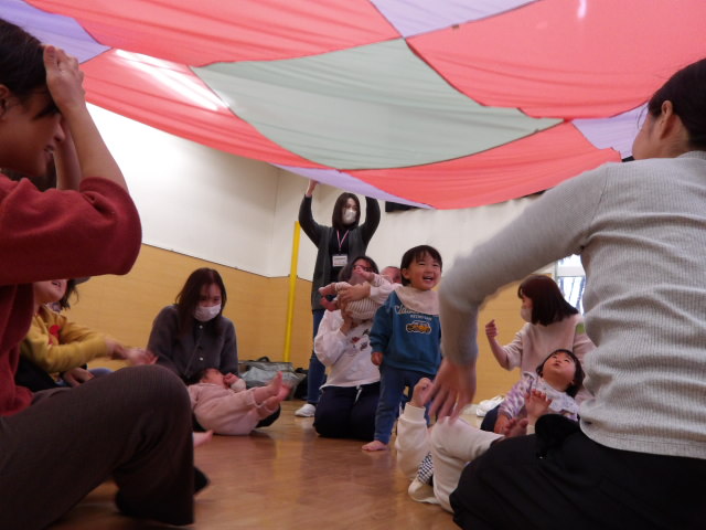 座っている幼児親子達に大きな布を上げ下げしている講師や職員の写真