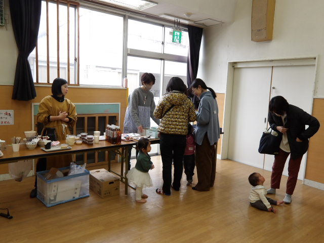 集会室にて、机に並べた平間銀座の商品を買う幼児親子3組と商品の用意をする職員2人の写真