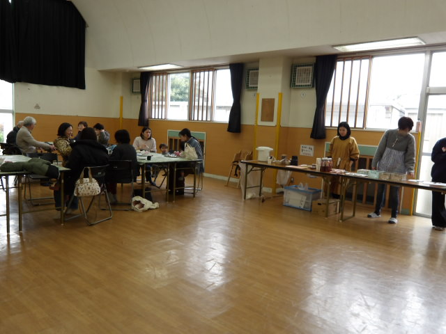 集会室で商品を販売する職員たちと、椅子に座りカフェを楽しみながら談笑する利用者たち