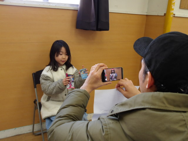 ポケモンカードを紹介する女子小学生とその様子をカンペを持ちながらスマホで撮影するお父さんの写真