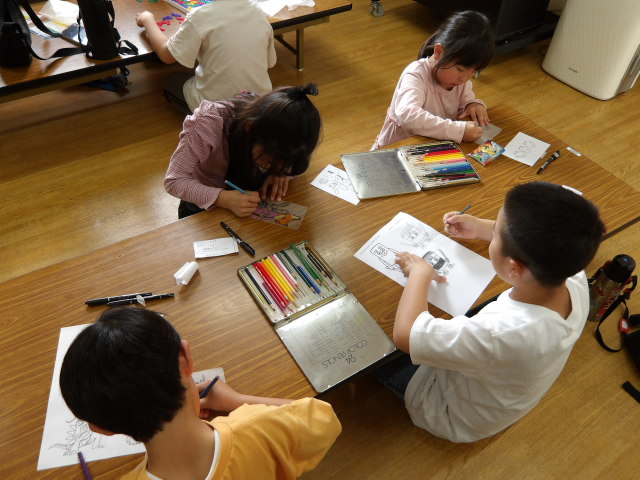 アニメのキャラクターが描かれたプラバンに色鉛筆で色を塗る小学生4人の写真