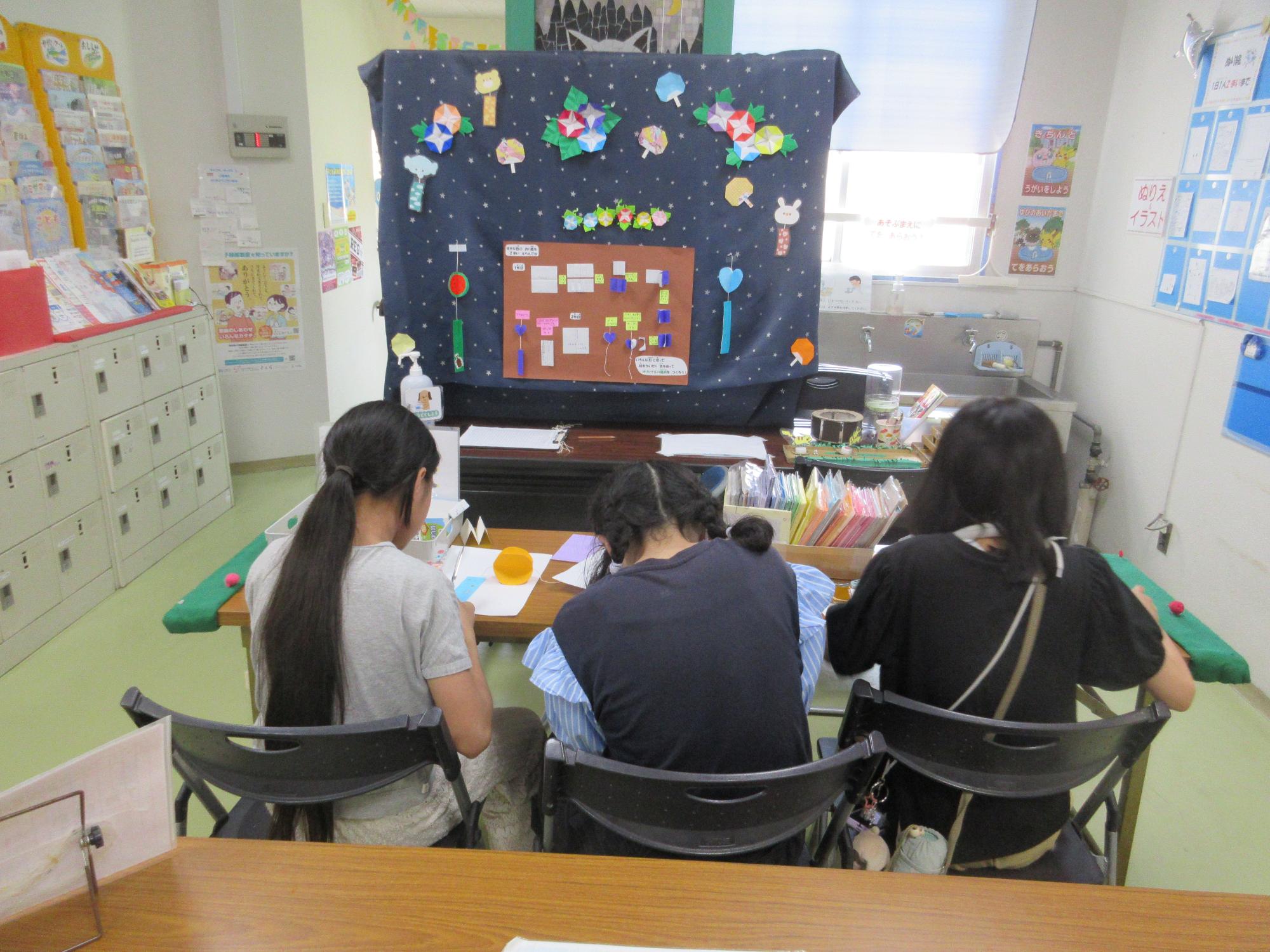 ホールには作り方が貼られておりテーブルに座り3人が折り紙で風鈴を作っているところ