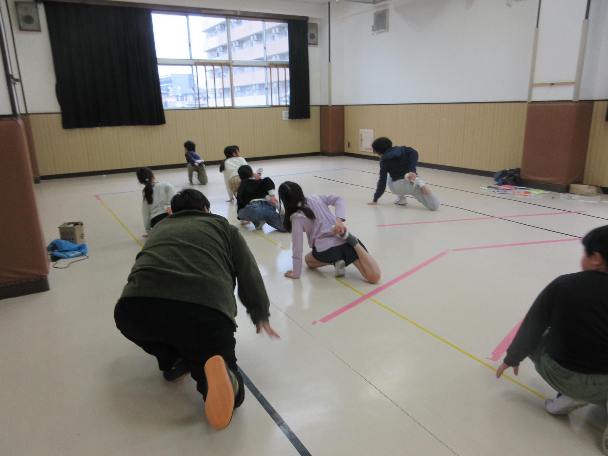 集会室で床にひざと手をついて座り片足を後ろに曲げ曲げた側の手で足首を持つストレッチをする子どもたち