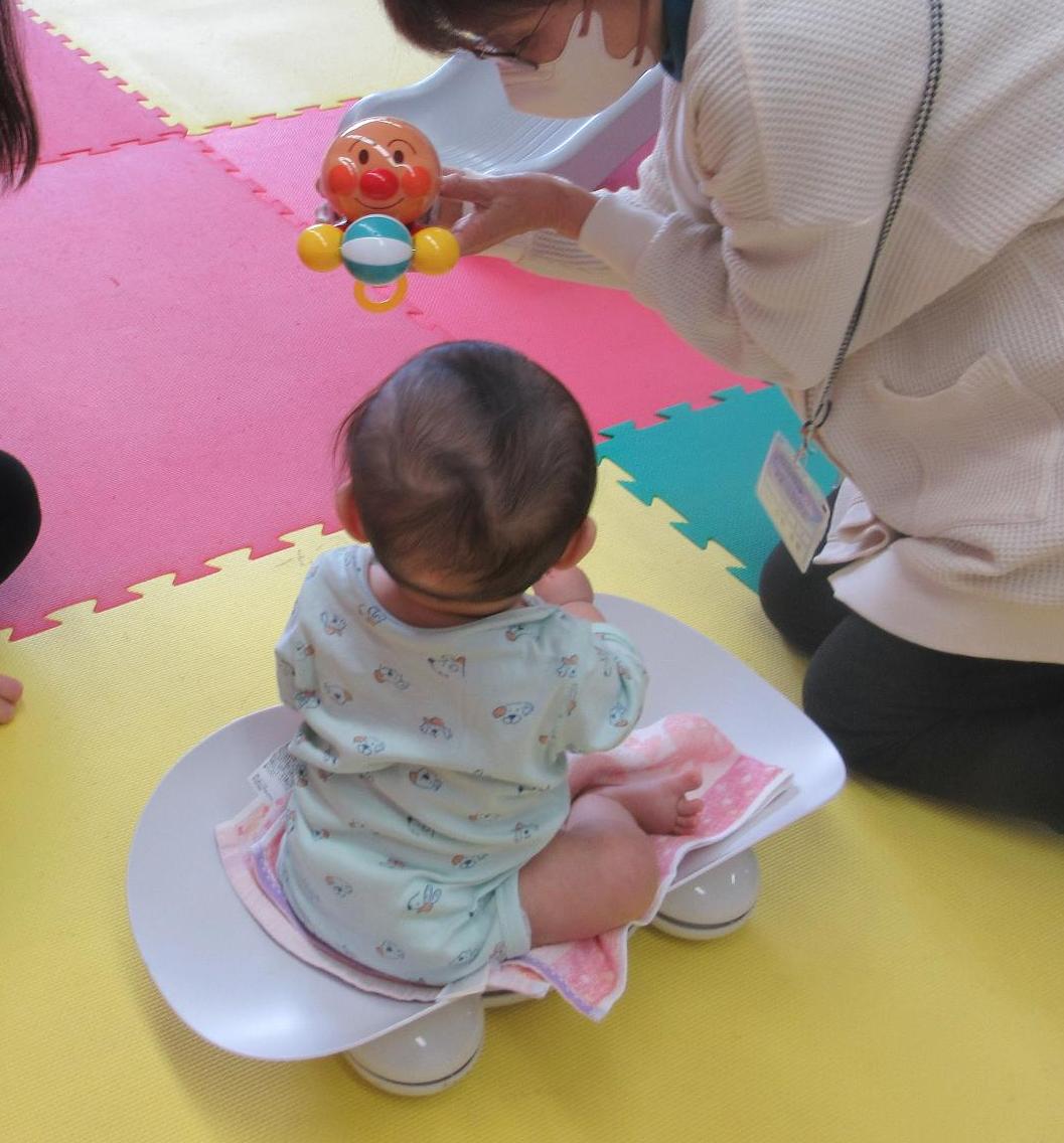 アンパンマンのおもちゃを眺めながらしっかりとお座りをして体重測定をする乳児さん