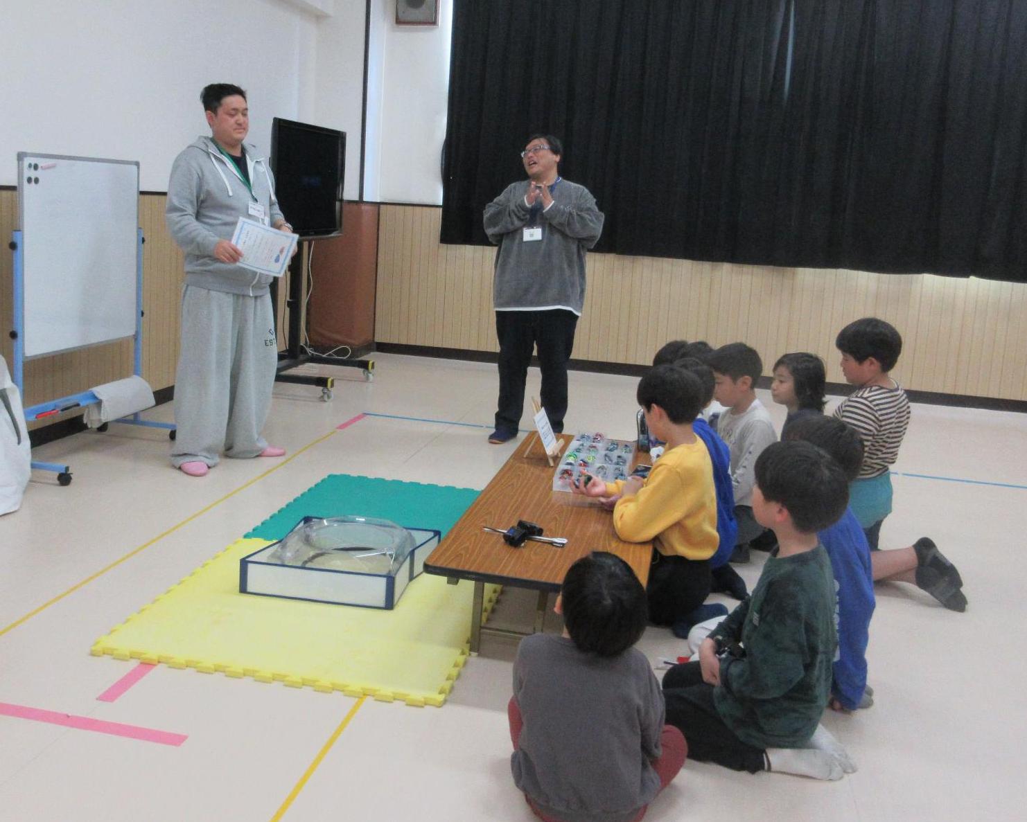 たくさんのベイブレードが入った箱がのったテーブルの前に座りベイブレードの講師から遊び方の説明を受ける子どもたち