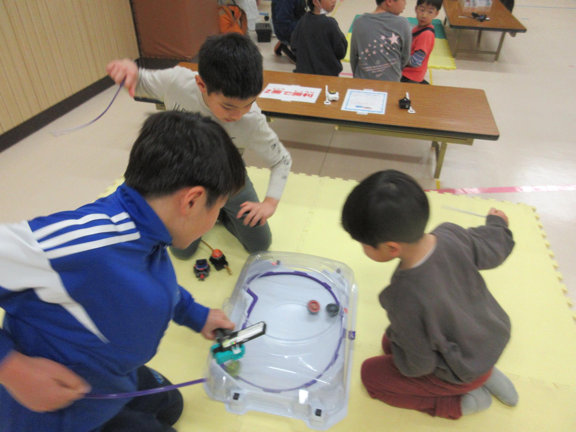 白い長方形の3人用のスタジアムでベイブレードのバトルをする子どもたち