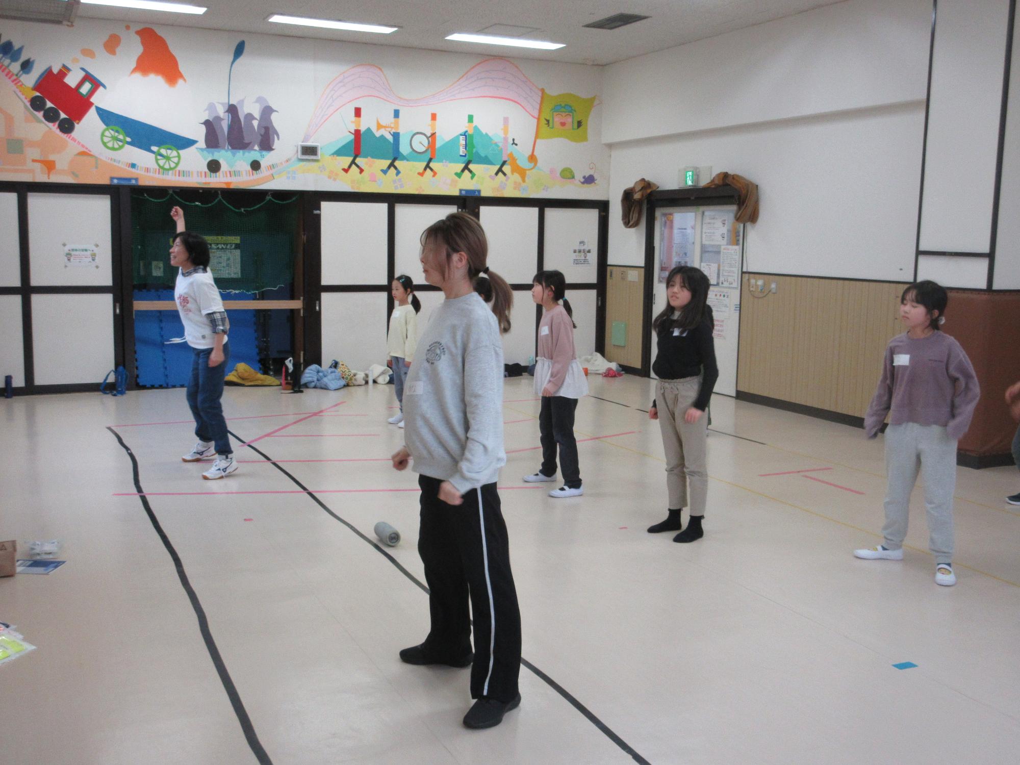 講師とアシスタントの後ろに二列に並び足を肩幅に広げ踊りを教わる子どもたち