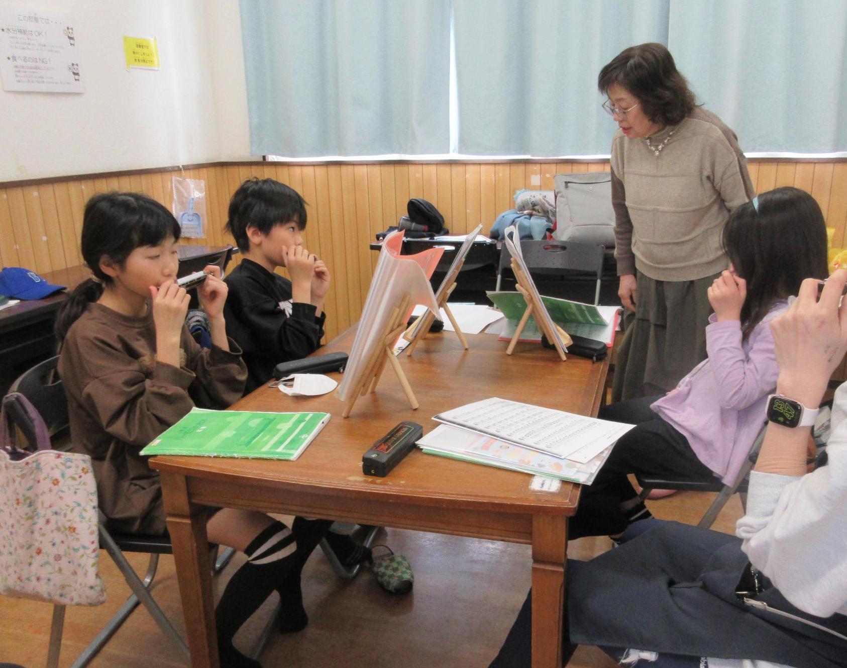 テーブルに楽譜を置き椅子に座りハーモニカを吹く子供たちと指導する先生