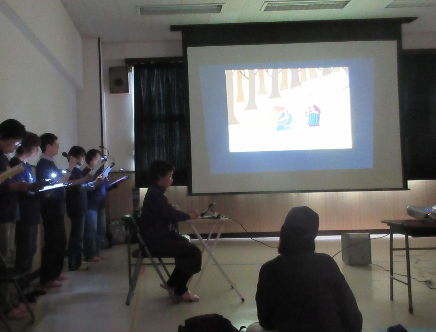電気を消した集会室で中央のスクリーンに物語の影絵を映し左壁に沿って演者が並び物語を読み上げる様子