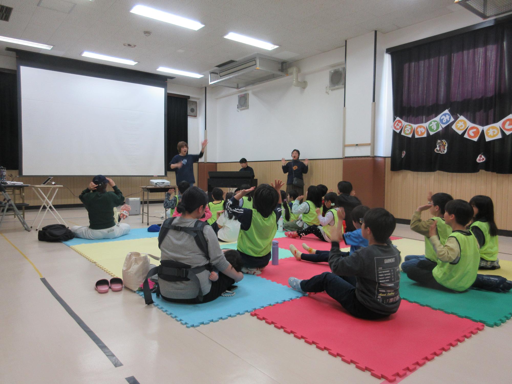 マットに座り手を口に当て手遊びを楽しむ観客たち