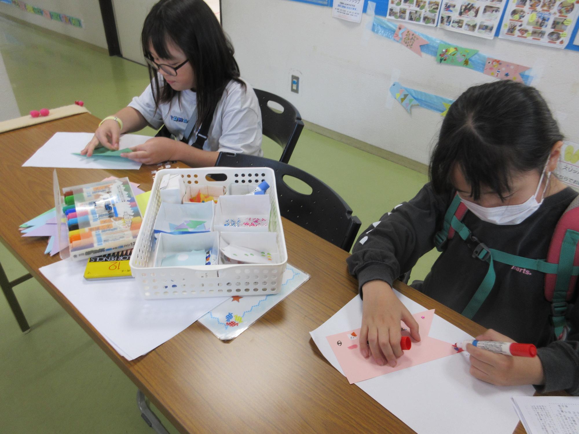 イスに座りテーブルでこいのぼり型の色紙に赤色のペンで鱗の絵を描く子どもとシールを貼る子ども