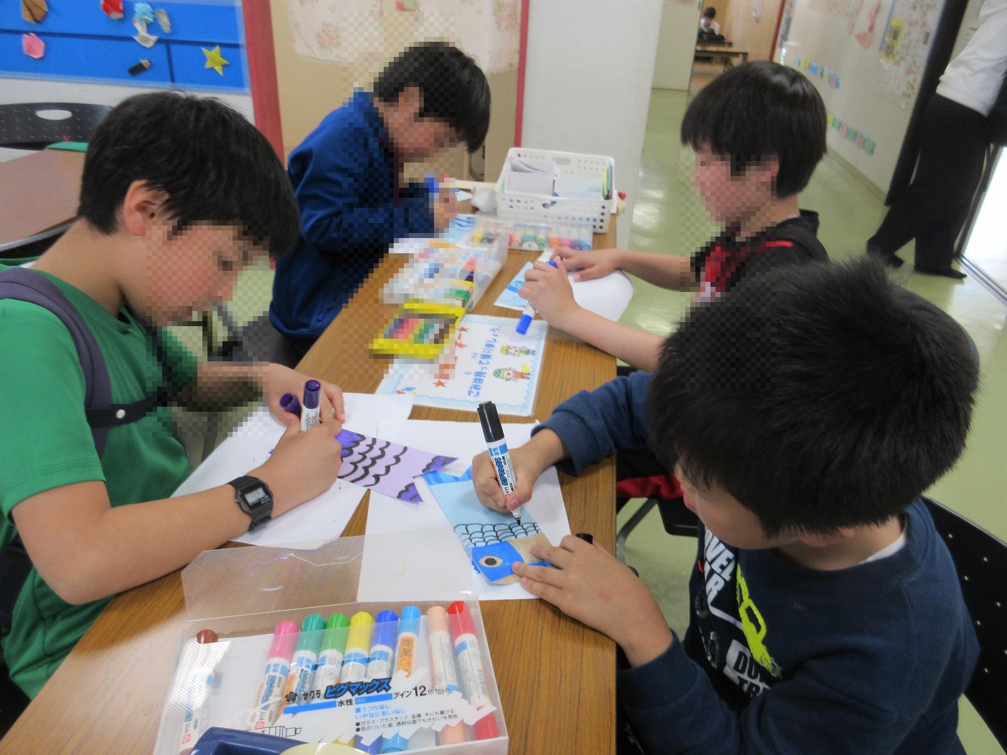 4人の小学生がイスに座りテーブルで鯉の形の色紙に黒と紫のペンで鱗を描いているところ