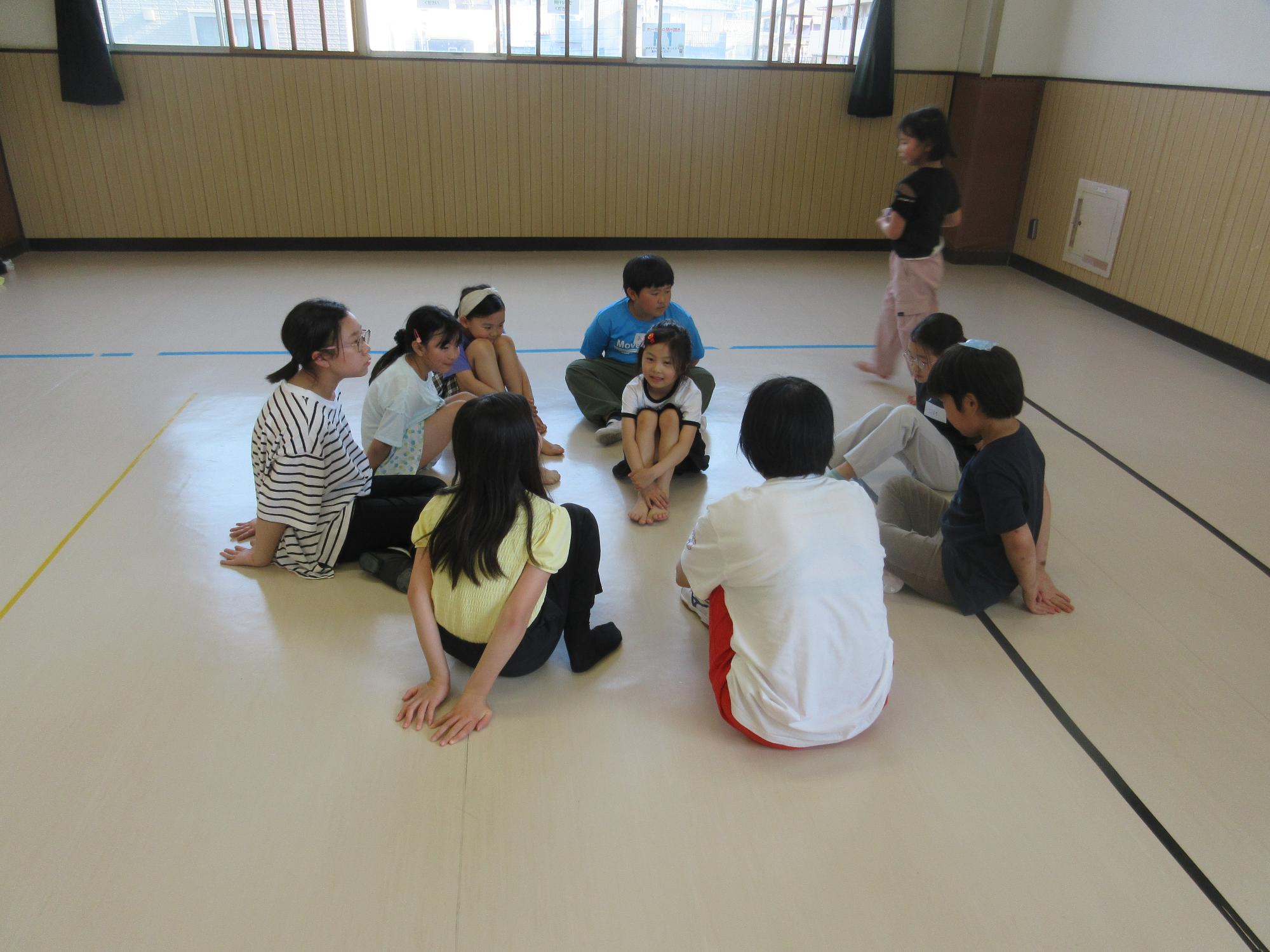 9人の子どもが輪になり内向きに座った外をハンカチを持った子供が次の鬼を誰にしようかと歩き回る様子
