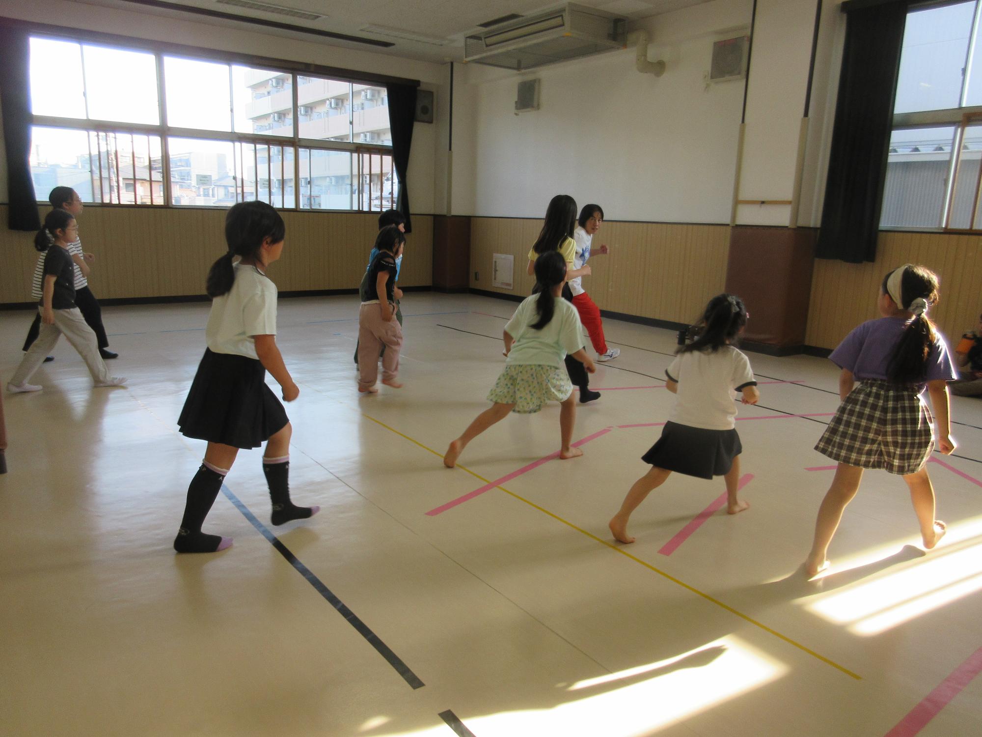 集会室に広がり、立って足を前後に開き前足を曲げるステップを練習する子どもたち