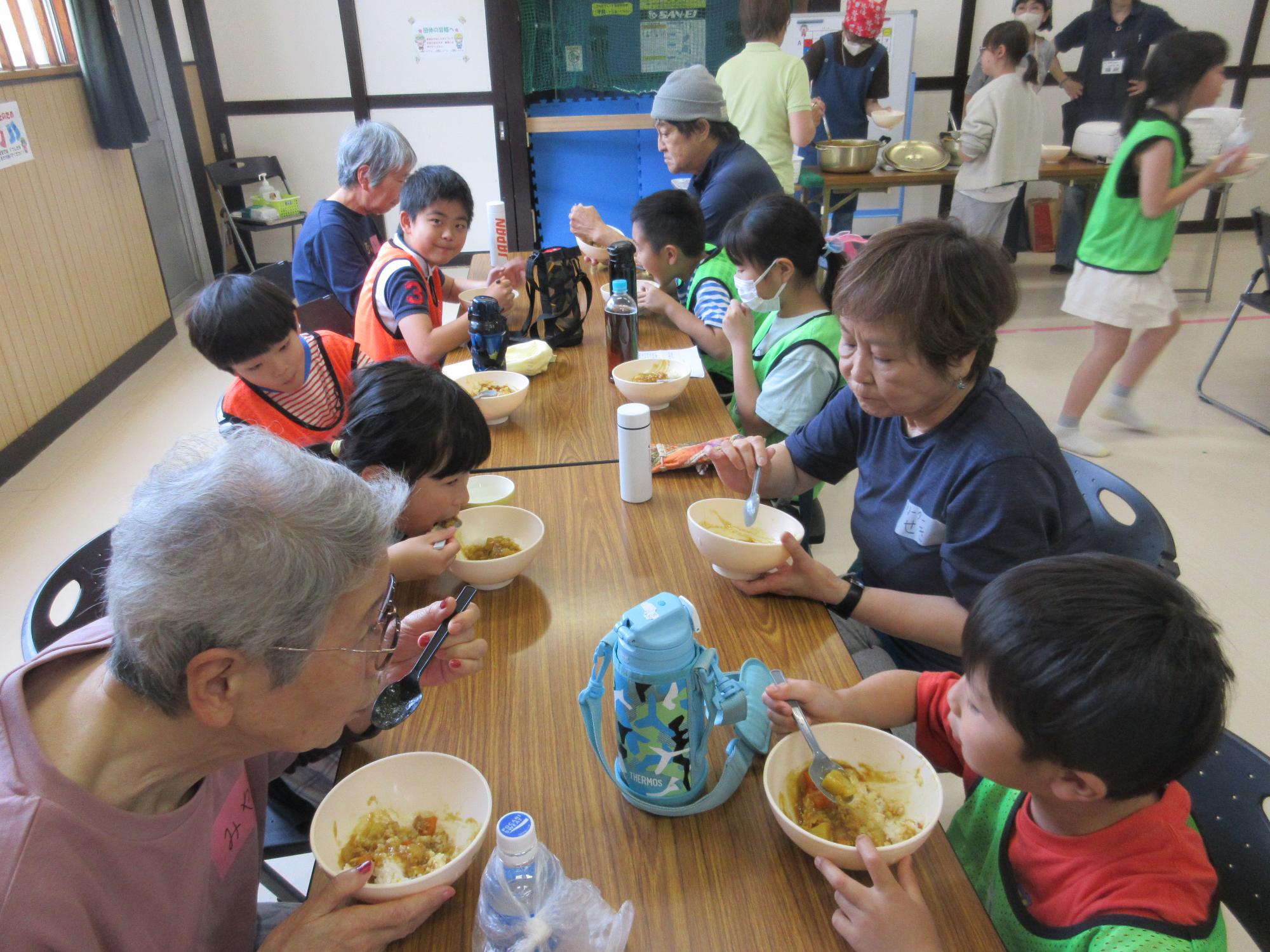 いこいの家の利用者と小学生が交互に座りテーブルで一緒にカレーライスを食べているところ