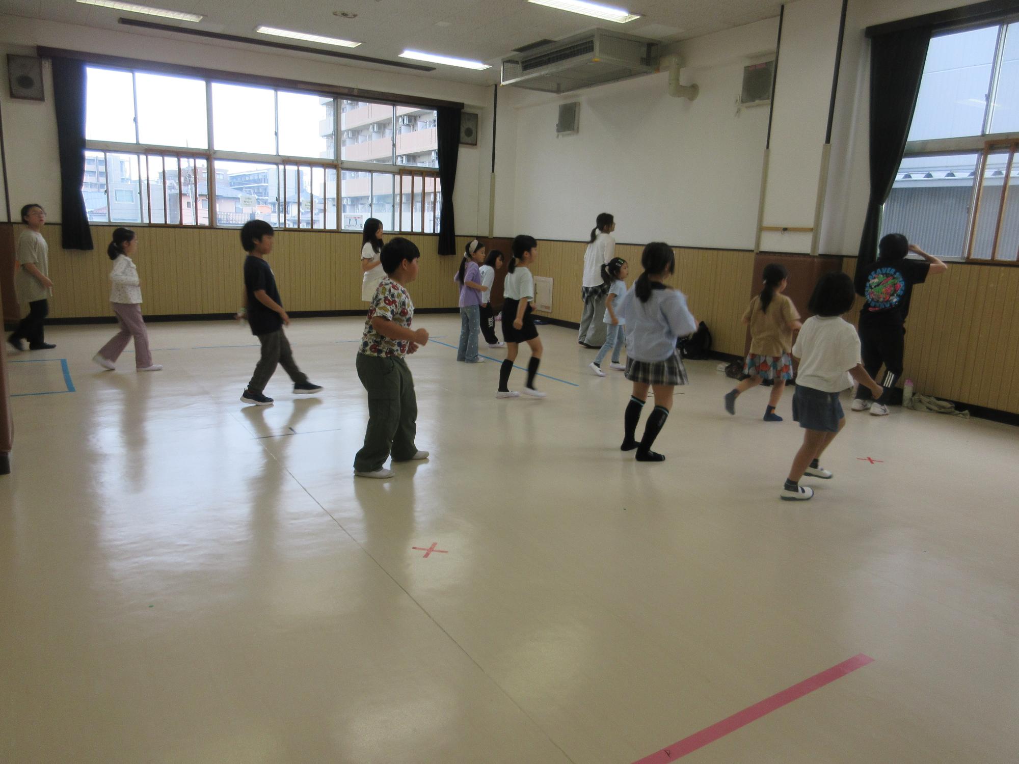 集会室で12人の子どもたちが先生の真似をして前後に足を出してステップをしているところ