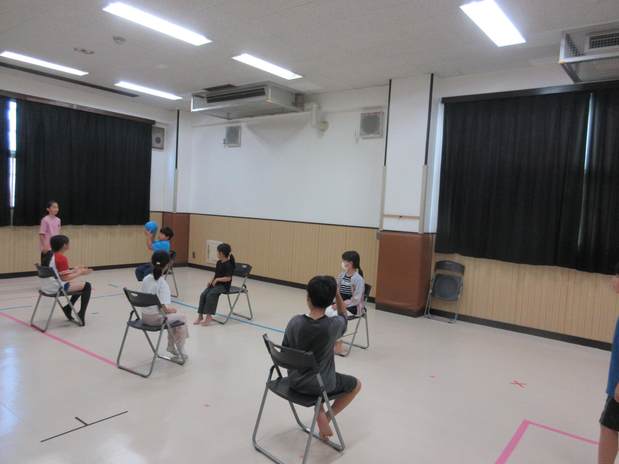 6人の子どもが向かい合わせに並んだいすにすわりはしのゴール係へボールをパスするところ
