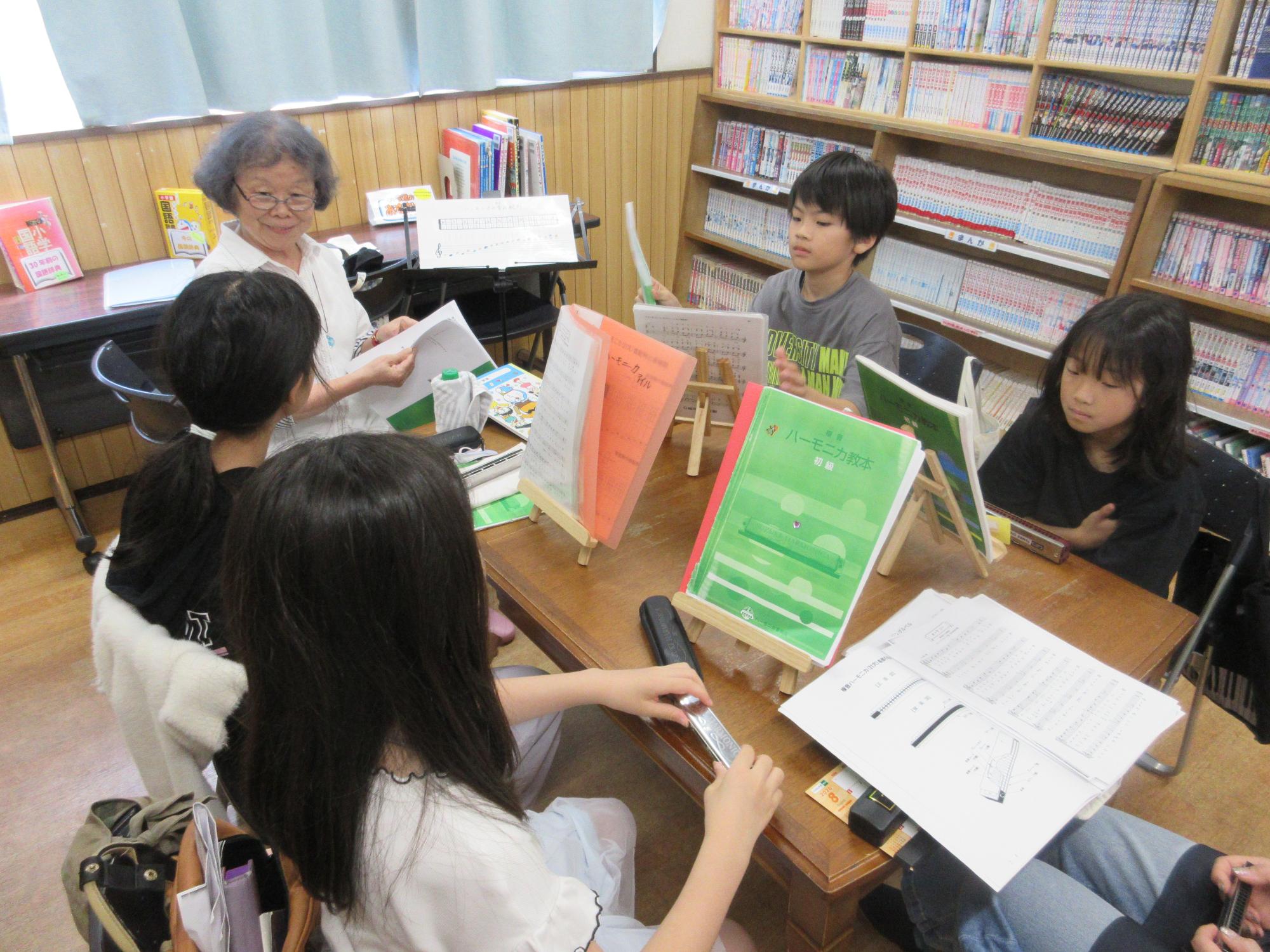 先生と5人の小学生がいすにすわりテーブルに楽譜をおいてハーモニカについて話を聞いているところ。