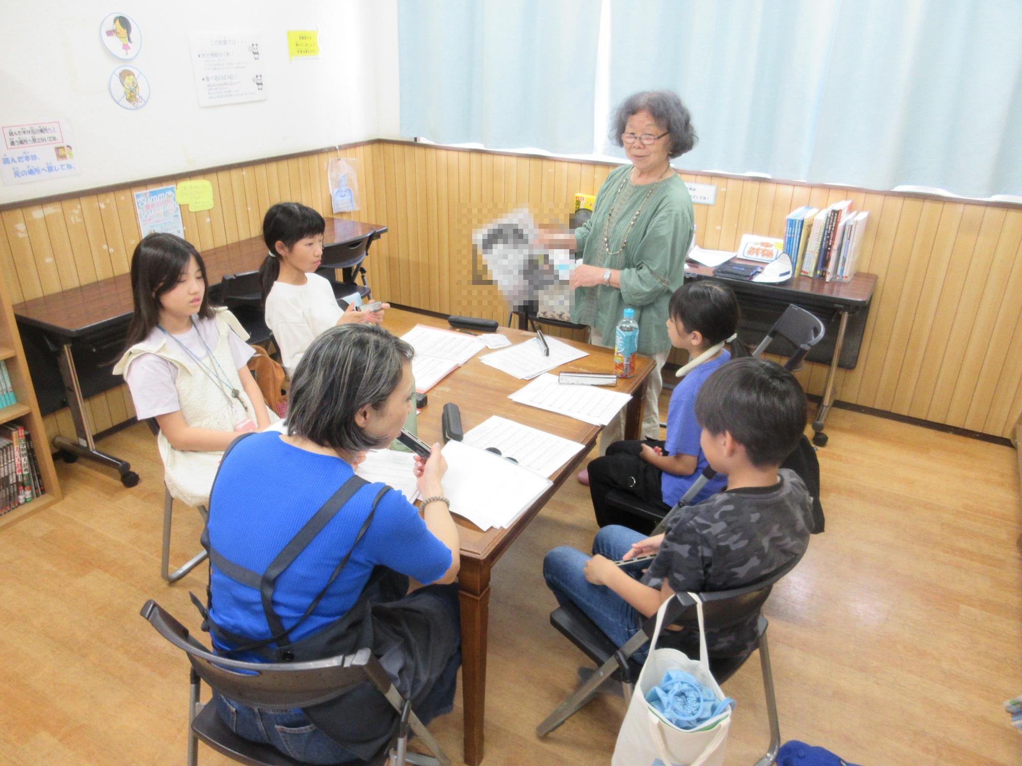 図書室で楽譜を広げたテーブルを囲み講師から4人の子どもと職員が教区の説明を受けているところ