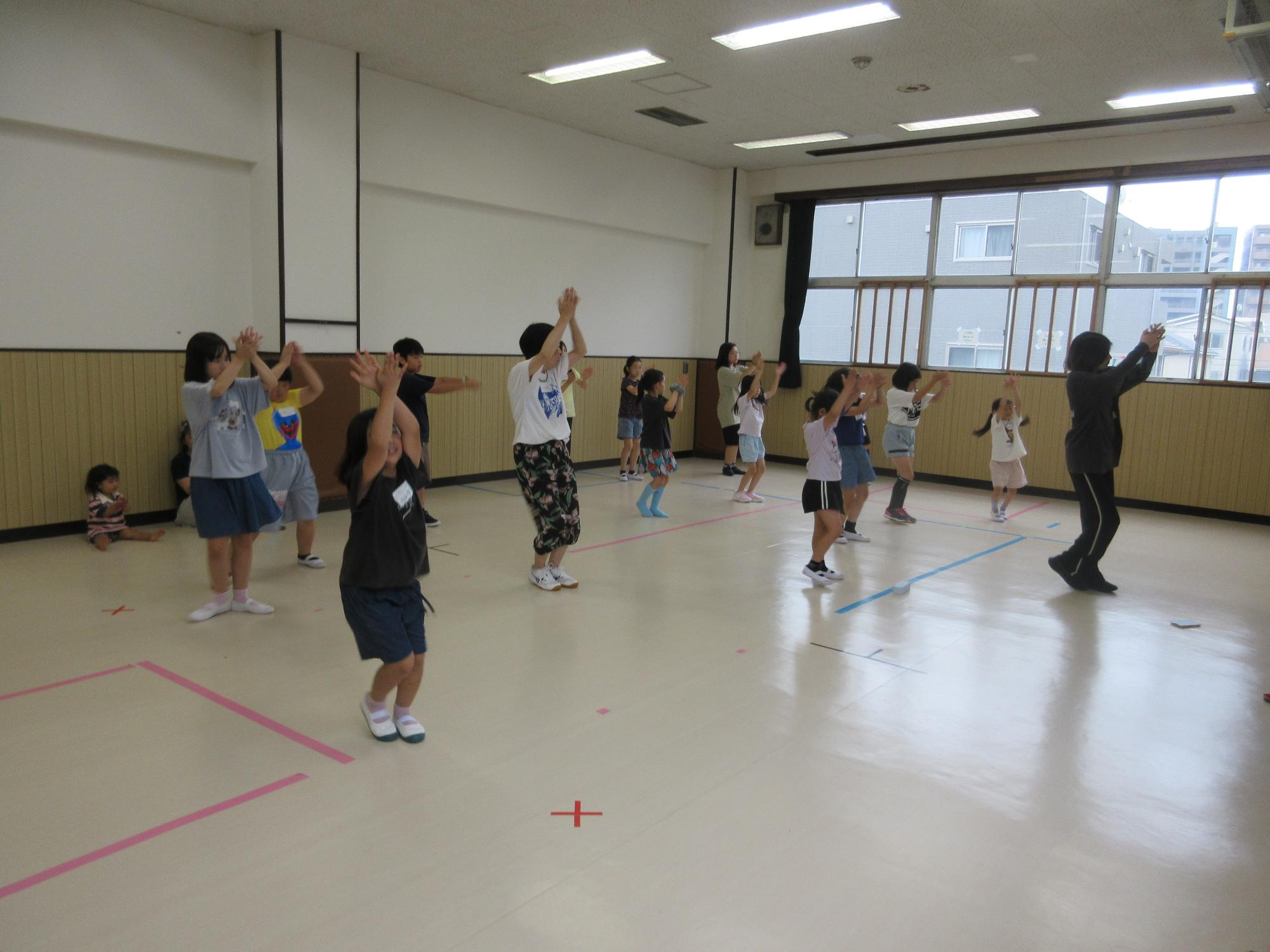 集会室で横3列に並び講師の手本をまねて頭の上で両手を合わせ両足を揃えてステップをしているところ