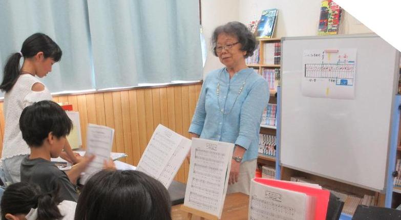ホワイトボードに楽譜をはり、楽譜の置かれたテーブルに座り先生のお話を聞く小学生