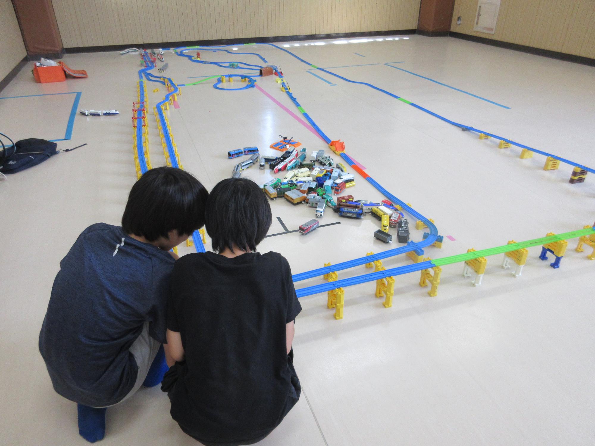 集会室に作ったながい長方形の線路を眺めどのように列車を走らせようかと相談する小学生