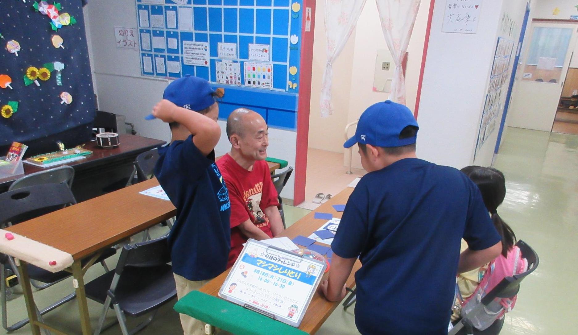 ホールで長テーブルをはさんで職員と小学生が座りこんちゃれに挑戦しようとしているところ