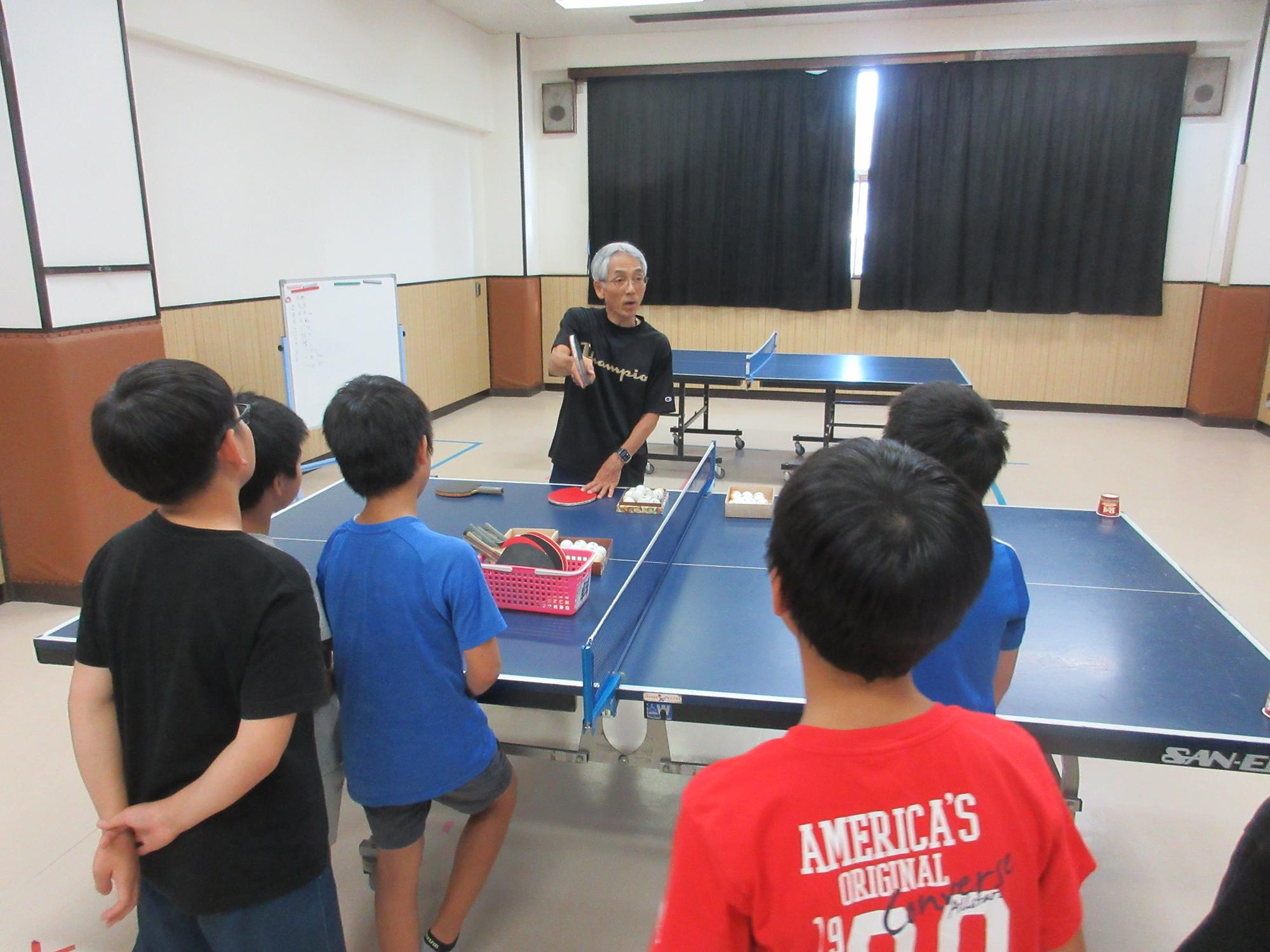 卓球台をはさみ講師が小学生にラケットの持ち方を教えているところ