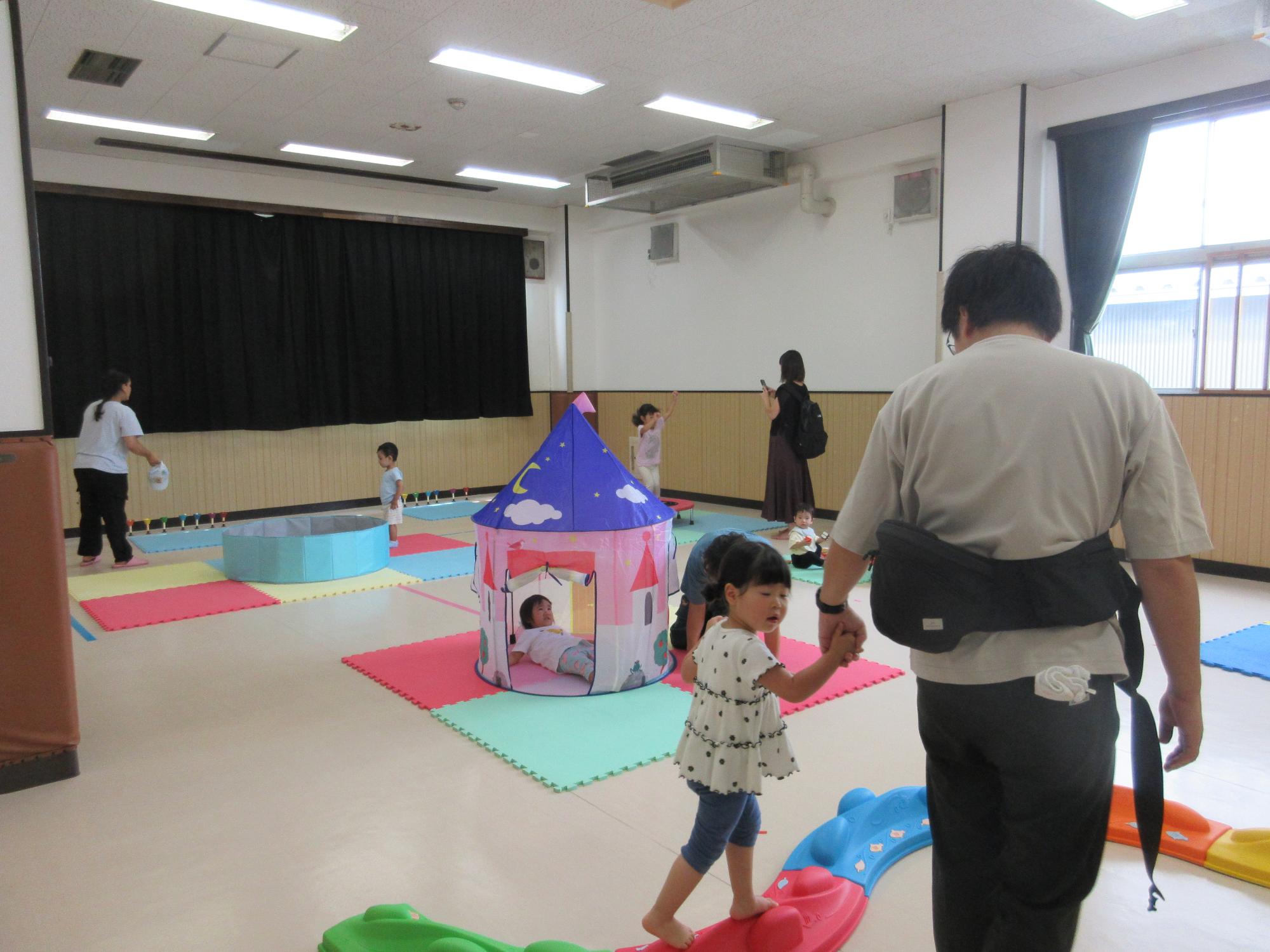 集会室に平均台とテントとボールプールとハンドベルとトランポリンがあり親子で遊んでいるところ