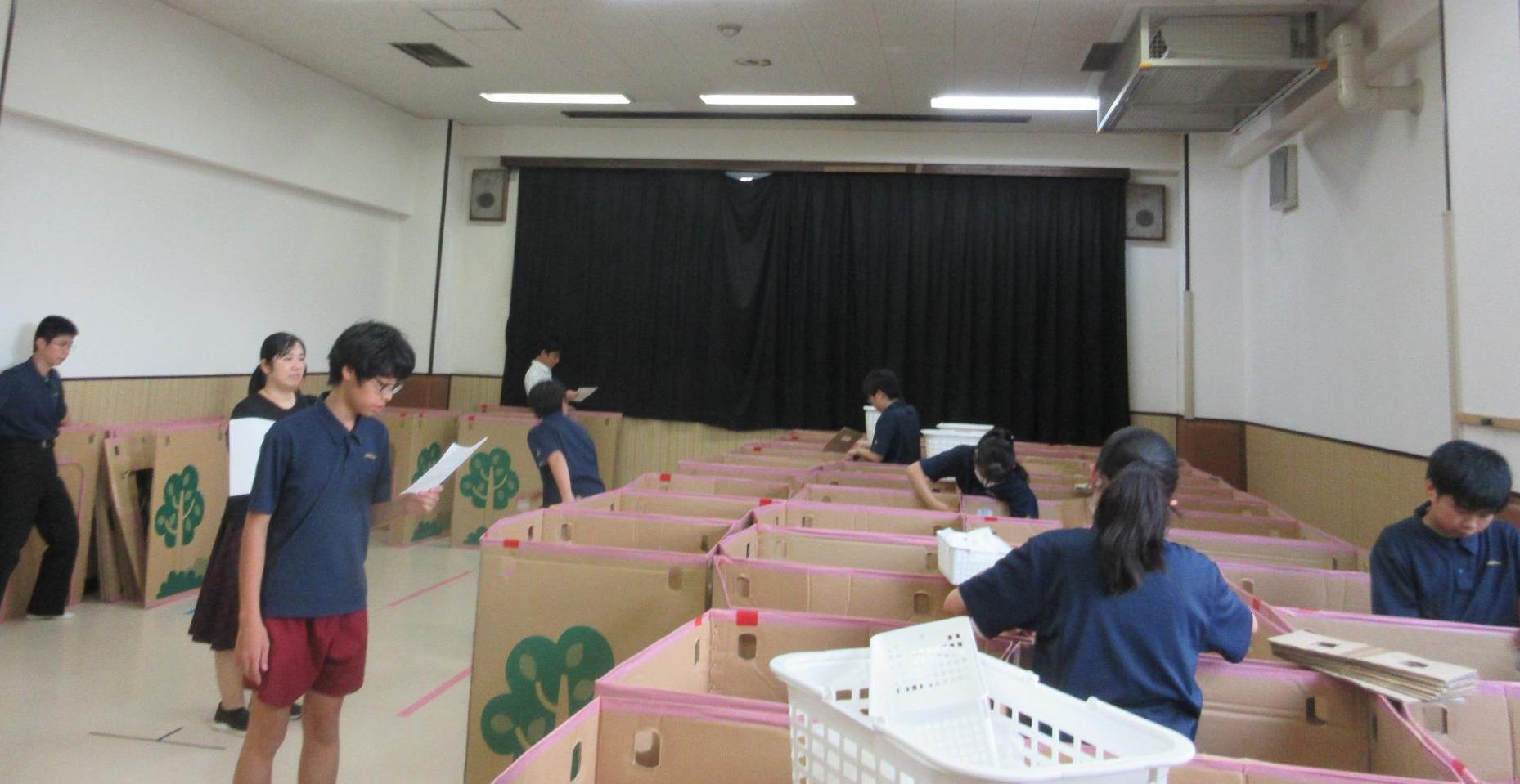 集会室の半分に四角く組み立てたダンボールを図面に合わせて並べる中学生