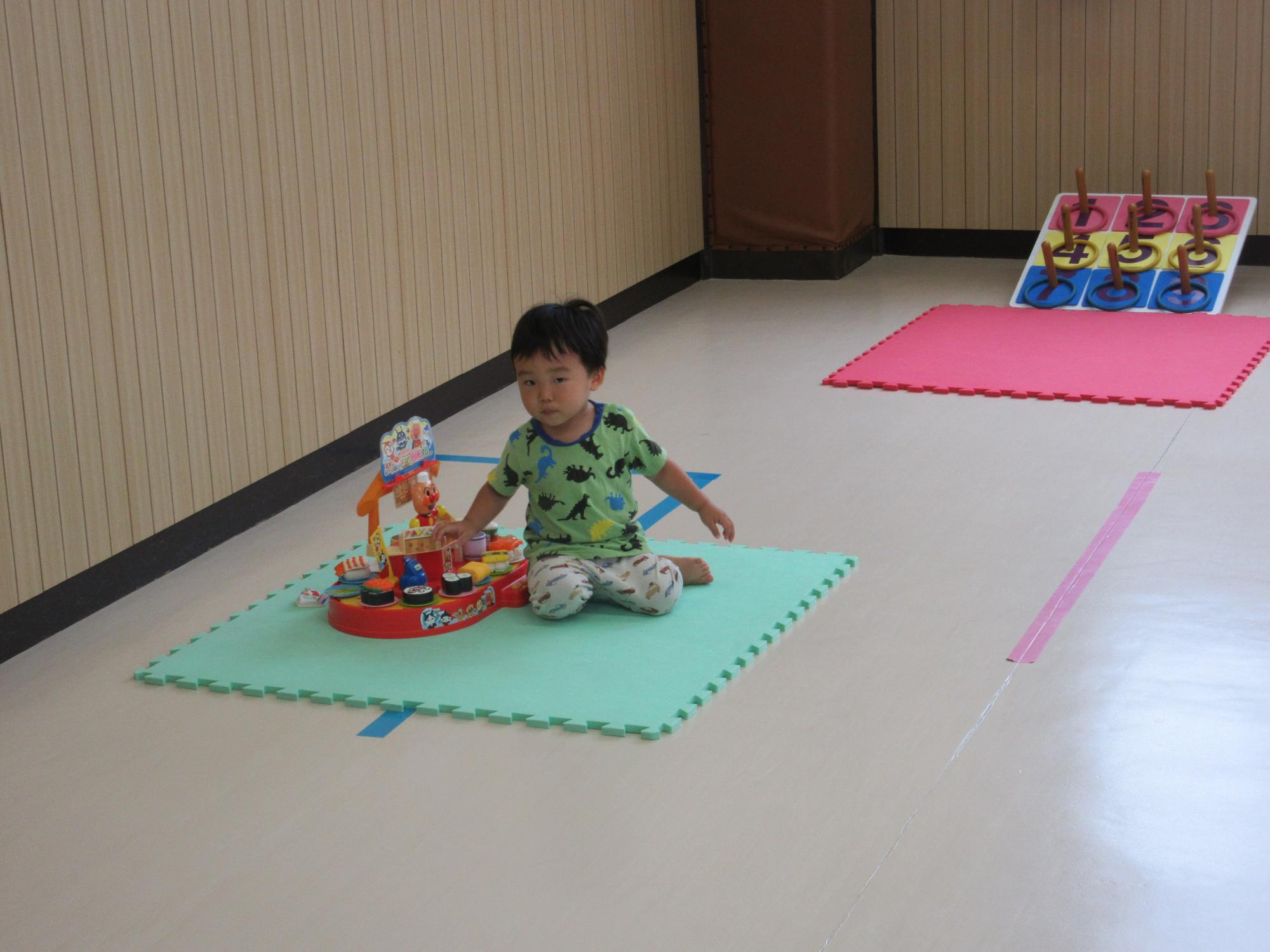 集会室に輪投げとアンパンマンのお寿司屋さんのおもちゃがありマットに座りお寿司屋さんで遊ぶ幼児
