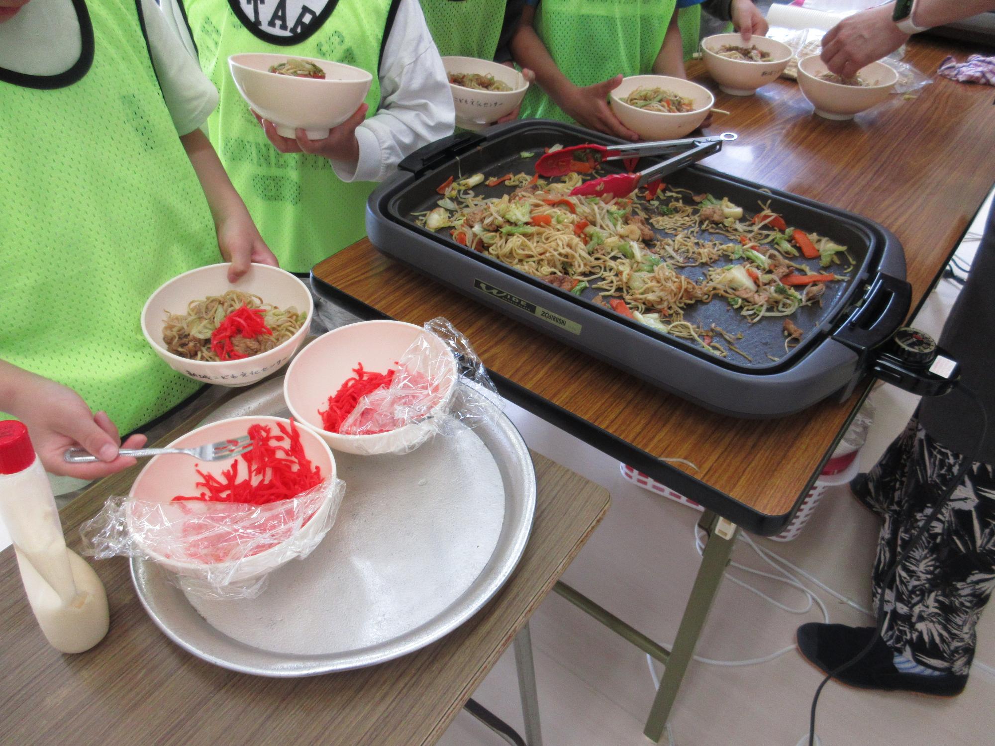 机にホットプレートに敷かれた焼きそばとトッピング用の紅しょうが並んでいる写真