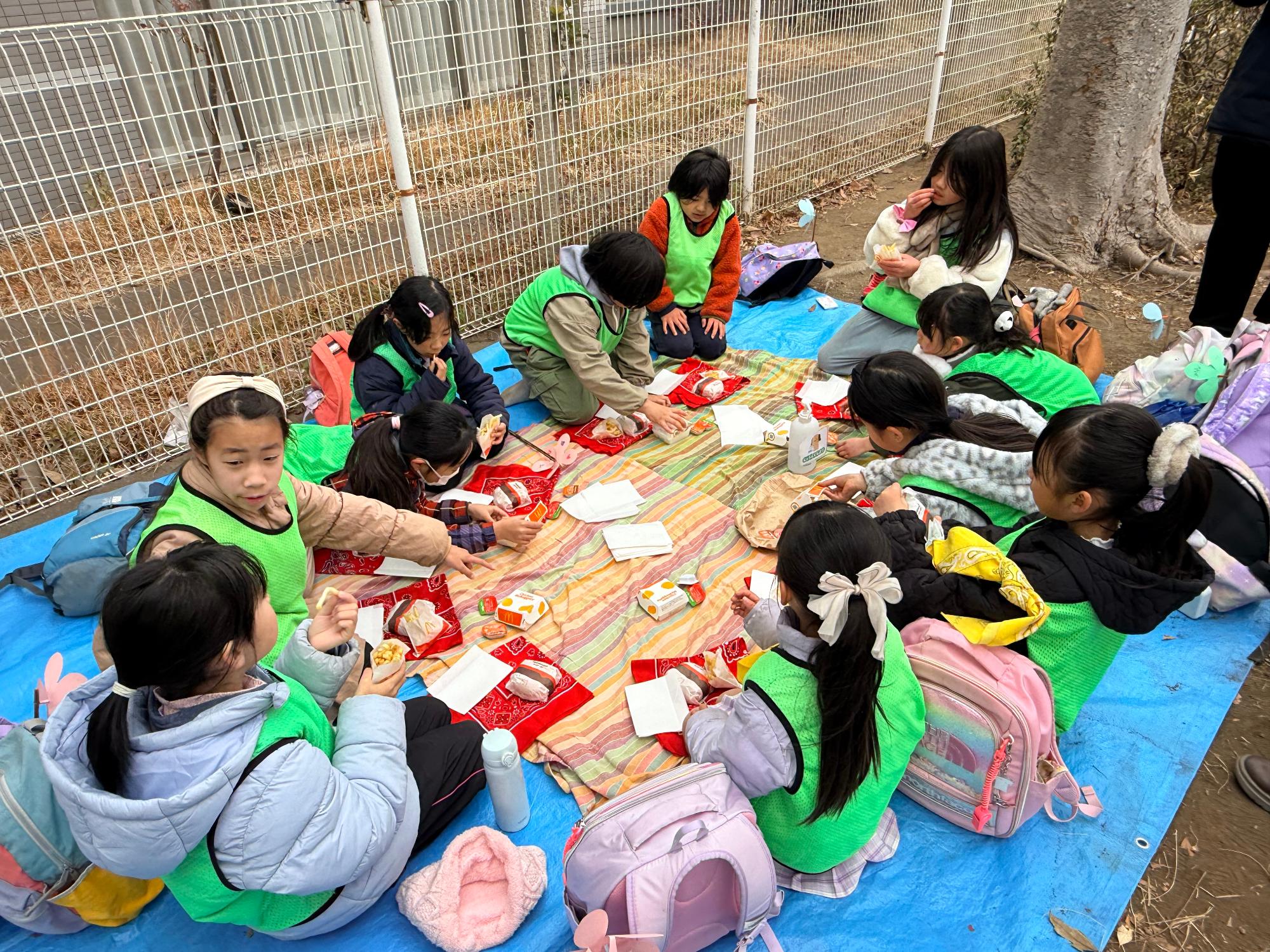 公園でブルーシートを敷きオレンジと緑色のストライプの布を広げ周りに子ども11人が座り赤いバンダナの上に乗せたハンバーガーとポテトとナゲットを食べる子どもたちに