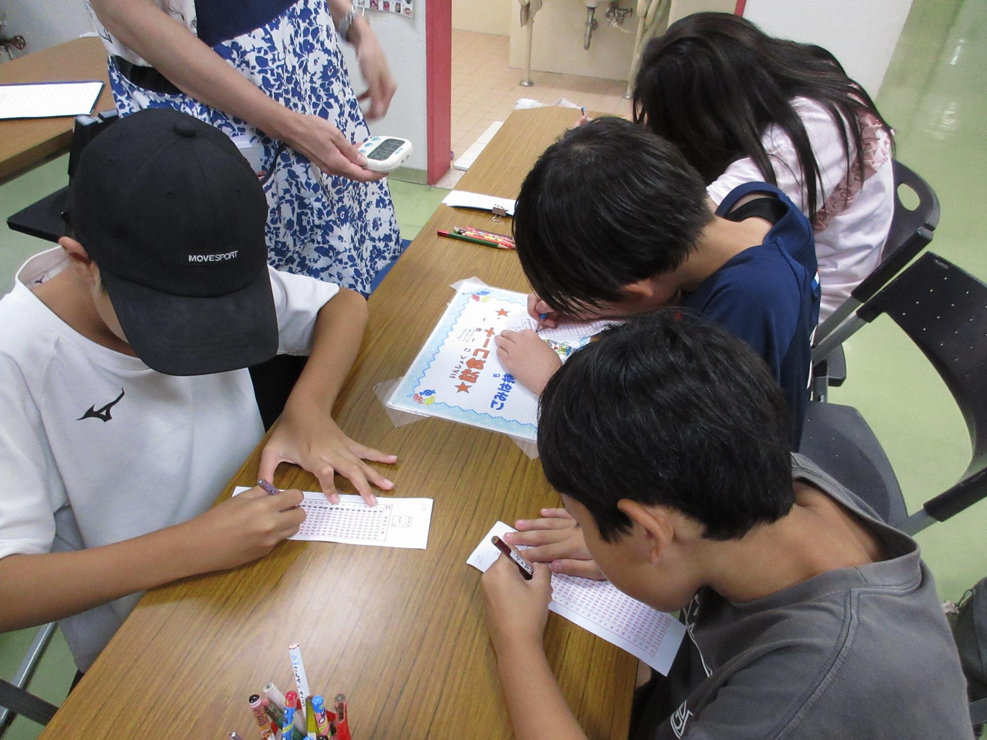 タイムを計っている職員とマークシートを塗りつぶす小学生4名の写真