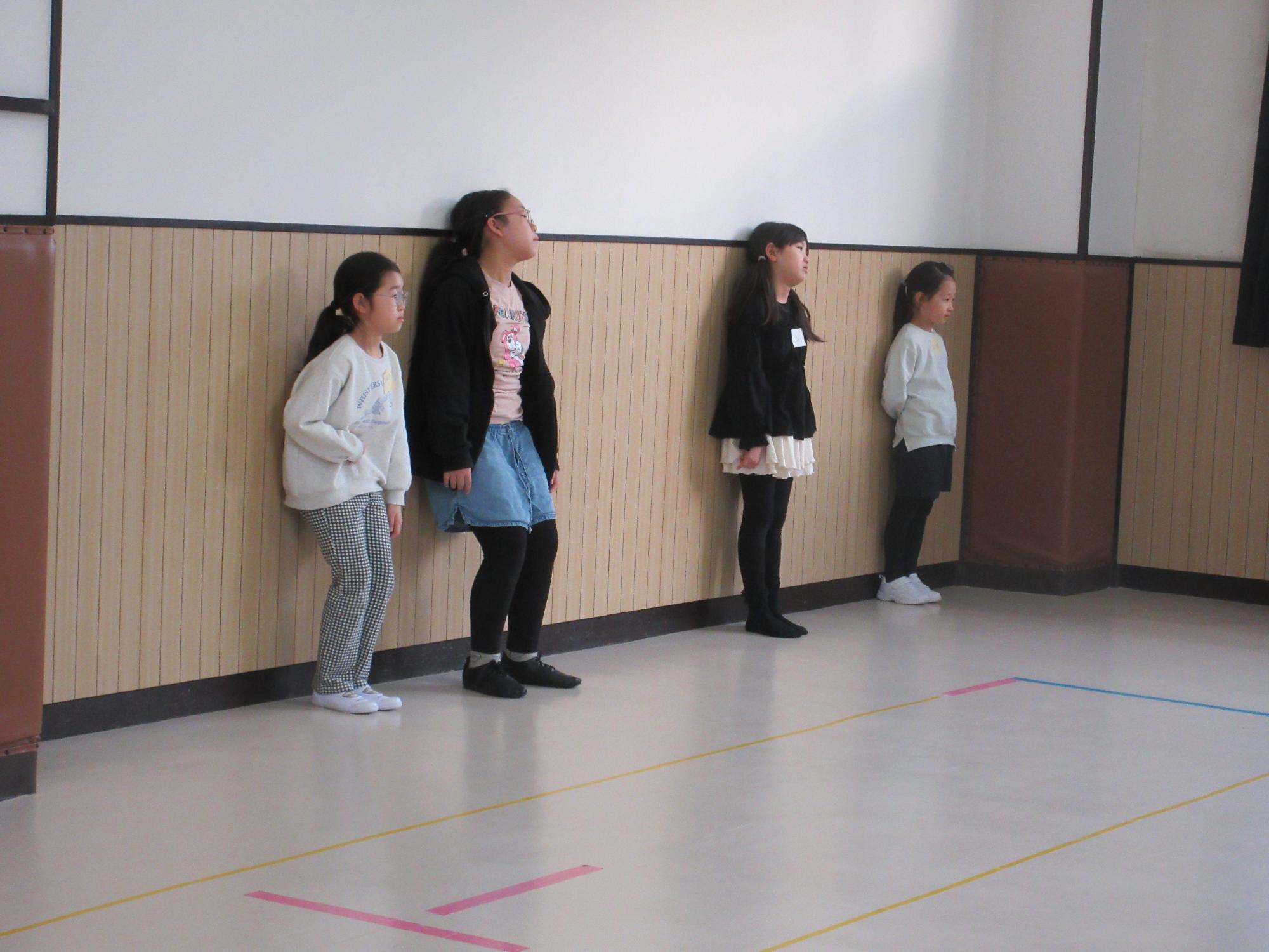 壁に背をつけてひざを曲げダンスのコツを学ぶ子どもたち