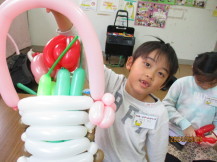 赤やピンク色の花が入った白色のカゴに付けた薄いピンク色の持ち手を持った男の子が、完成した花かごをカメラに向かって見せている写真