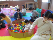 カラフルな色のボールが入った円形のボールプールに、赤ちゃんや2名の女の子が入り遊んでいる写真