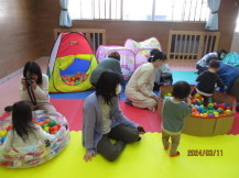 円形の段ボール製とビニール製の2つのボールプールで遊んでいる子供を、母親が近くで見たり写真を撮ったりして楽しんでいる写真