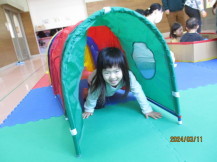 室内に設置されたジョイントマットの上に赤や緑色のトンネルで遊んだ女の子が出てくる様子の写真