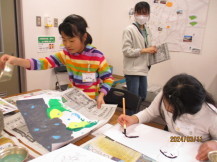 下書きした紙しばいの絵が描かれた画用紙に絵の具で色を塗っている2名の女の子の写真