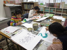 新聞紙を敷いたテーブルの席に座った3名の女の子が、絵の具を使って紙芝居を製作している様子の写真