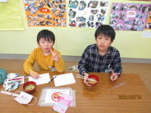 長机に並んで座った2名の男の子が赤色のお椀に入った大根のお味噌汁を食べ、カメラに向かってピースサインをしている写真写真