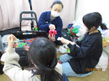 青色の服を着た女性講師に教わりながらバルーンアートを作っている2名の女の子の写真