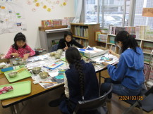 2台の長机をくっつけた上に新聞紙を敷き、椅子に座った女性と3名の女の子が紙しばいの絵に色を塗っている様子の写真